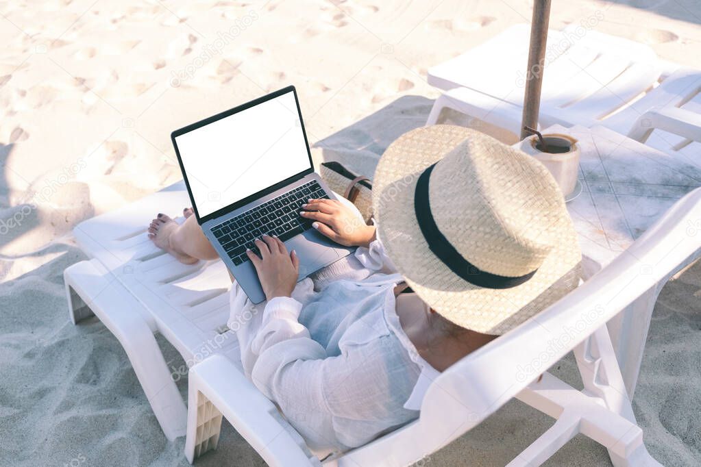 Top view mockup image of a woman holding and using laptop computer with blank desktop screen while laying down on beach chair on the beach
