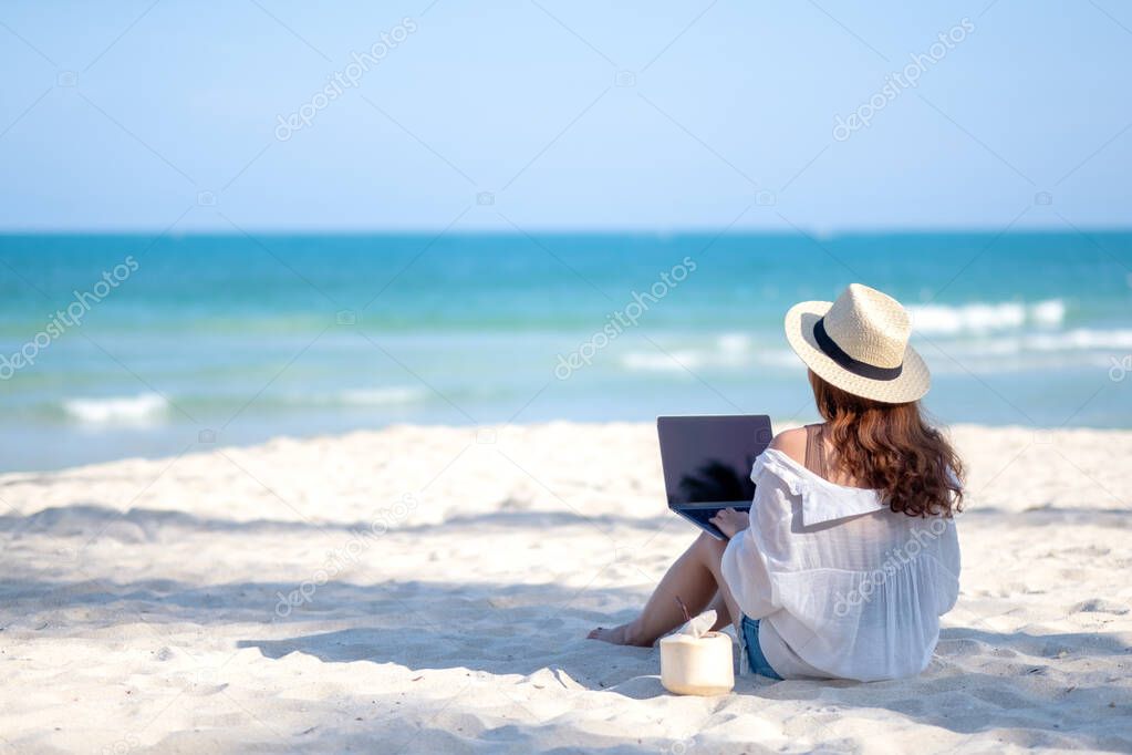 A woman using and typing on laptop computer keyboard while sitting on a beautiful beach