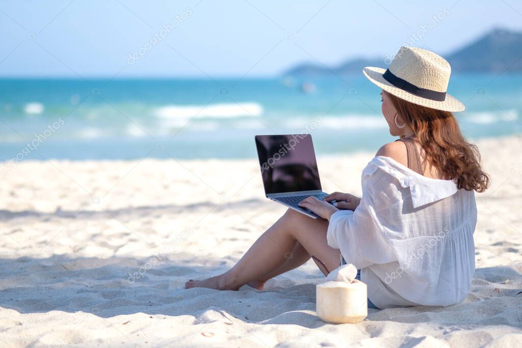 A woman using and typing on laptop computer keyboard while sitting on a beautiful beach