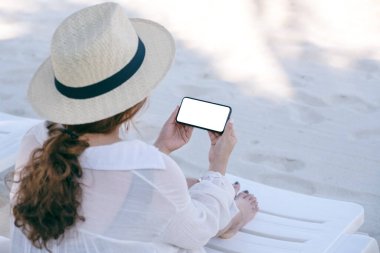 Plaj sandalyesinde otururken elinde boş masaüstü ekran olan beyaz bir cep telefonu tutan bir kadının görüntüsü.