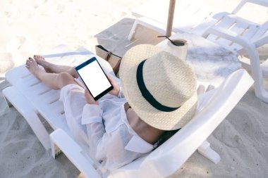 Sahilde plaj sandalyesinde uzanırken elinde boş masaüstü ekran tableti olan bir kadının en üstteki görüntüsü.