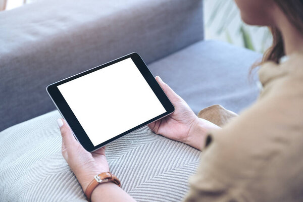 Mockup image of a woman holding black tablet pc with blank white desktop screen horizontally while sitting in living room with feeling relaxed