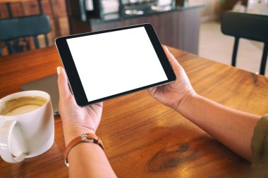 Ellerin siyah tableti tutuş görüntüsü. Boş beyaz ekran ve ahşap masada kahve fincanı var. 