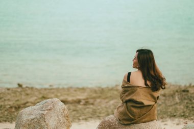 Güzel bir Asyalı kadın deniz kıyısındaki kayada oturmaktan zevk alır.