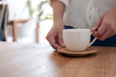 Kafe 'de beyaz bir fincan sıcak kahveyi ahşap masada tutan bir kadının yakın plan görüntüsü.
