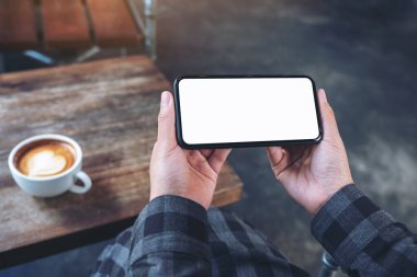 Kafe 'deki ahşap masada, boş ekranlı, beyaz cep telefonu tutan ellerin görüntüsü.