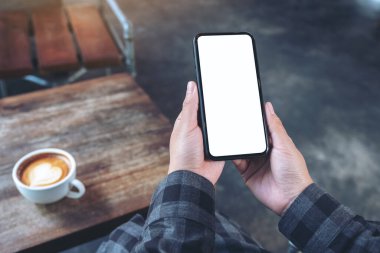 Kafedeki ahşap masada kahve fincanı ve boş ekranlı beyaz cep telefonu tutan ellerin görüntüsü.