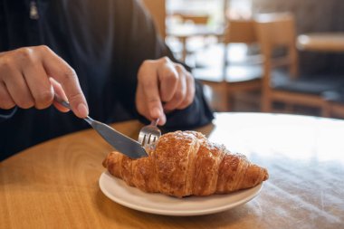 Yakından bakınca, ahşap masada kahvaltı için çatal ve bıçakla kruvasan kesen eller görülüyor. 