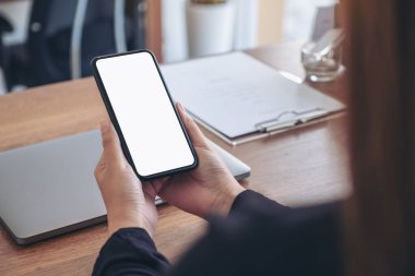 Ofisteki ahşap masadaki dizüstü bilgisayarlı, boş beyaz ekranlı, siyah cep telefonlu ellerin görüntüsü.