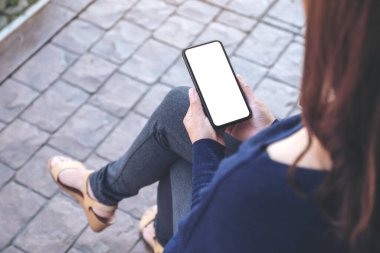 Açık havada otururken elinde beyaz ekranlı siyah bir cep telefonu tutan bir kadının görüntüsü.