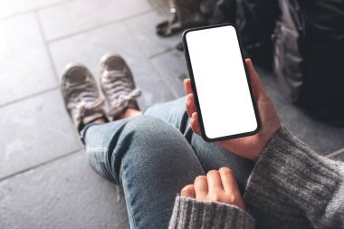 Üst görünümde, yerde bavulla otururken elinde boş masaüstü ekran olan siyah bir cep telefonu tutan bir kadının görüntüsü.