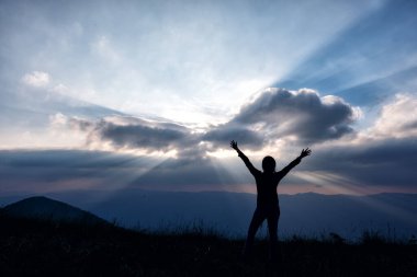 Silüet bir kadın görüntüsü ayakta ve el kaldırıyor, akşamları dağların manzarasıyla gün batımını seyrediyor.