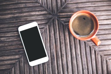 Beyaz bir cep telefonunun, boş siyah masaüstü ekran görüntüsü ve eski ahşap masa arkaplanındaki bir fincan kahvenin üst görünümü.