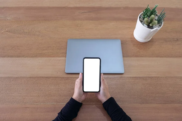 オフィスの木製テーブルの上に空白の白い画面の携帯電話とラップトップを保持している手のモックアップイメージのトップ表示 — ストック写真