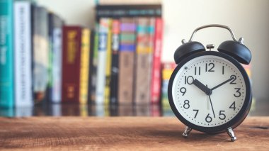 Arkaplanda bulanık kitaplık bulunan ahşap masadaki siyah alarmlı saatin yakın plan resmi