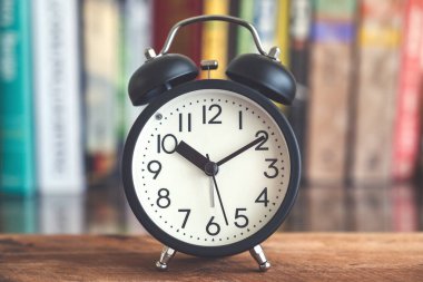 Arkaplanda bulanık kitaplık bulunan ahşap masadaki siyah alarmlı saatin yakın plan resmi