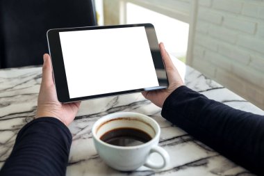 Siyah tabletli, beyaz ekranlı ve kafedeki kahve fincanının olduğu ellerin görüntüsü. 