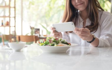 Kafedeki beyaz tabakta salata çekmek için akıllı telefon kullanan Asyalı bir kadının yakın plan görüntüsü.