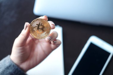 Ofisinde dizüstü bilgisayarı ve cep telefonu olan altın renkli bir Bitcoin tutan bir iş kadını resmi.