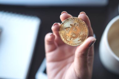 Ofisinde dizüstü bilgisayarı ve cep telefonu olan altın renkli bir Bitcoin tutan bir iş kadını resmi.