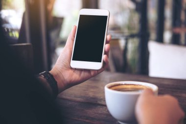 Ellerin beyaz cep telefonu, boş siyah masaüstü ekran ve eski kafenin ahşap masasında bir kahve fincanı tutarken görüntüsü.