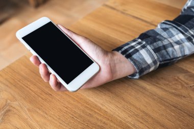 Kafedeki ahşap masada boş siyah masaüstü ekranı olan beyaz cep telefonunu tutan el resmi.