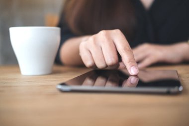 Kafedeki ahşap masanın üzerinde beyaz kahve fincanı olan tableti işaret eden, dokunan ve kullanan bir kadının yakın plan görüntüsü.