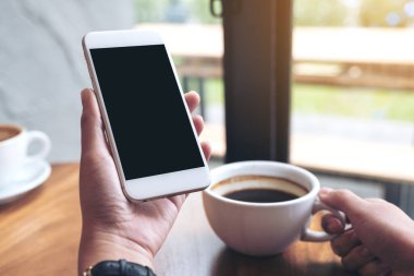 Kafede kahve içerken beyaz cep telefonunu siyah ekranla tutan ellerin görüntüsü.