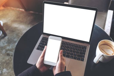 Masada boş beyaz masaüstü ekranı olan dizüstü bilgisayarı kullanırken boş cep telefonunu tutan ellerin en üstteki görüntüsü 