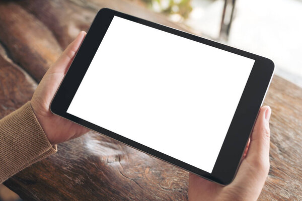 Mockup image of hands holding black tablet pc with blank white desktop screen on wooden table