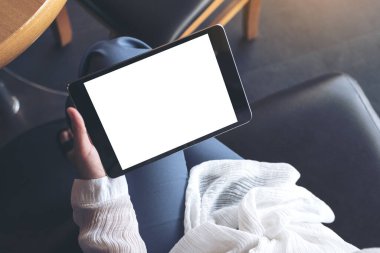 Üst görünümde çapraz bacaklı oturan ve elinde boş beyaz masaüstü ekranlı siyah tablet bilgisayar tutan bir kadının görüntüsü 