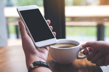 Kafede kahve içerken beyaz cep telefonunu siyah ekranla tutan ellerin görüntüsü.