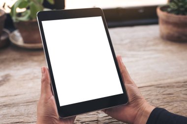 Eski ahşap masa üzerinde boş beyaz ekranlı siyah tableti tutan ellerin görüntüsü. 