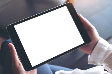 Üst görünümde çapraz bacaklı oturan ve elinde boş beyaz masaüstü ekranlı siyah tablet bilgisayar tutan bir kadının görüntüsü 