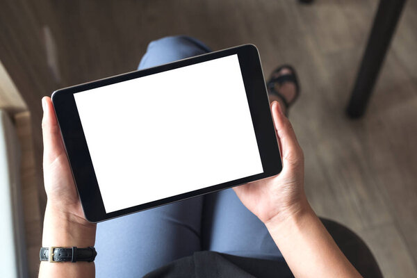 Top view mockup image of a woman sitting cross legged and holding black tablet pc with blank white desktop screen in office