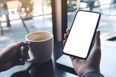 Kafedeki masada kahve fincanı ve dizüstü bilgisayarı olan, boş beyaz ekranlı, siyah cep telefonlu bir kadının el resmi.