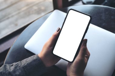 Kafedeki masada dizüstü bilgisayarı olan, boş beyaz masaüstü ekranlı, elinde siyah cep telefonu olan bir kadının görüntüsü.