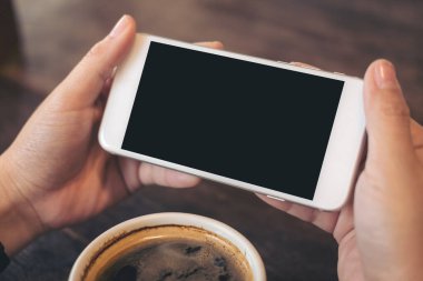 Kafedeki ahşap masada bir fincan kahveyle oyun oynayıp izlemek için beyaz cep telefonu ve boş siyah ekran tutan ellerin görüntüsü.