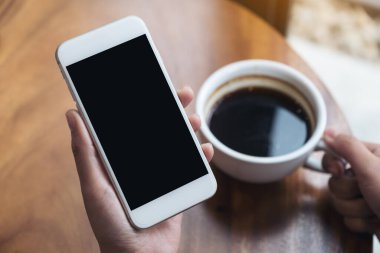 Kafede kahve içerken beyaz cep telefonlu, siyah ekranlı bir kadının ellerinin görüntüsü.