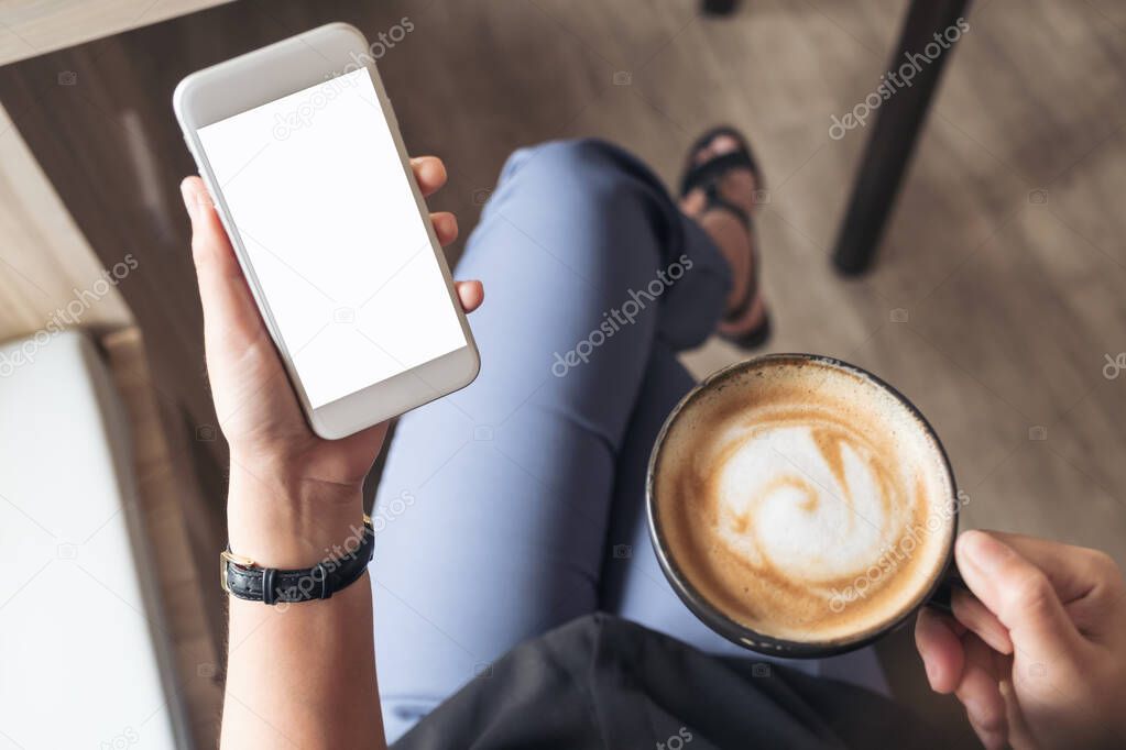 Top view mockup image of a woman's hand holding white mobile phone with blank desktop screen while drinking coffee in cafe