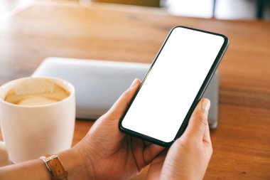 Masasında laptop ve kahve fincanı olan ve elinde boş masaüstü ekranı olan siyah bir cep telefonu tutan bir kadının görüntüsü.