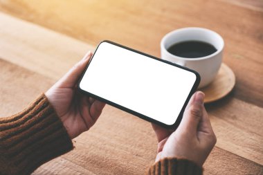 Elinde siyah bir cep telefonu olan bir kadının resmi. Yatay olarak boş ekran ve ahşap masada kahve fincanı var.