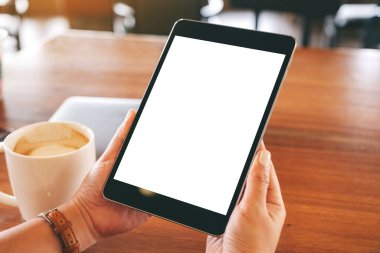 Siyah tabletli, beyaz ekranlı ve ahşap masada kahve fincanı olan ellerin görüntüsü. 