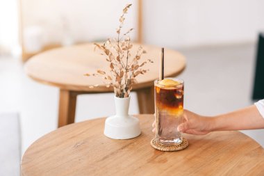 Yakından bakınca, minimumda bir kafede ahşap masada bir bardak buzlu hindistan cevizi kahvesi tutan bir el görülüyor.