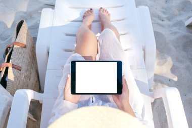 Sahildeki plaj sandalyesinde uzanırken elinde siyah bir tablet bilgisayar kullanan bir kadının en üstteki görüntüsü.