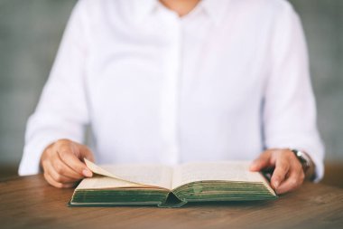 Ahşap masada elinde kitap tutan ve okuyan bir kadının yakın plan resmi.
