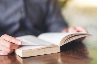 Ahşap masada elinde kitap tutan ve okuyan bir kadının yakın plan resmi.