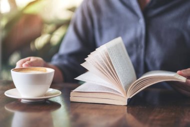 Tahta masada kahve içerken elinde kitap tutan ve okuyan bir kadının yakın plan resmi.