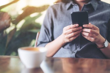 Kafe 'de elinde kahve fincanıyla cep telefonu tutan ve kullanan bir kadının yakın plan görüntüsü.