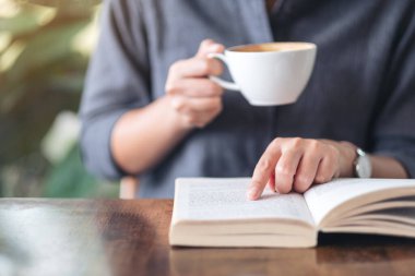 Ahşap masada kahve içerken kitap okuyup işaret eden bir kadının yakın plan resmi.